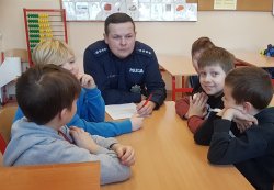 Zdjęcie przedstawia umundurowanego policjanta i pięciu chłopców. Siedzą oni przy szkolnym stoliku. Policjant tłumaczy zasady prawidłowego alarmowania.
