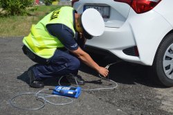 Umundurowany policjant Wydziału Ruchu Drogowego do układu wydechowego białego samochodu  podłącza urządzenie służące do pomiaru emisji spalin