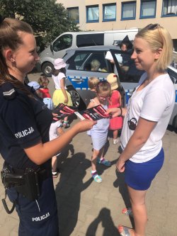 umundurowana policjantka wręcza kobiecie ulotkę profilaktyczną, za nimi widoczna jest grupa dzieci i policyjny oznakowany radiowóz