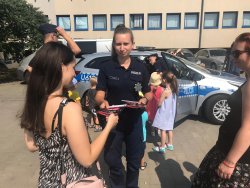 umundurowana policjantka wręcza kobiecie ulotkę profilaktyczną, za nimi widoczna jest grupa dzieci i policyjny oznakowany radiowóz