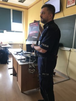 Policjant prowadzi prelekcję w klasie szkolnej, trzymając plakat dotyczący kampanii Narkotyki i dopalacze zabijają